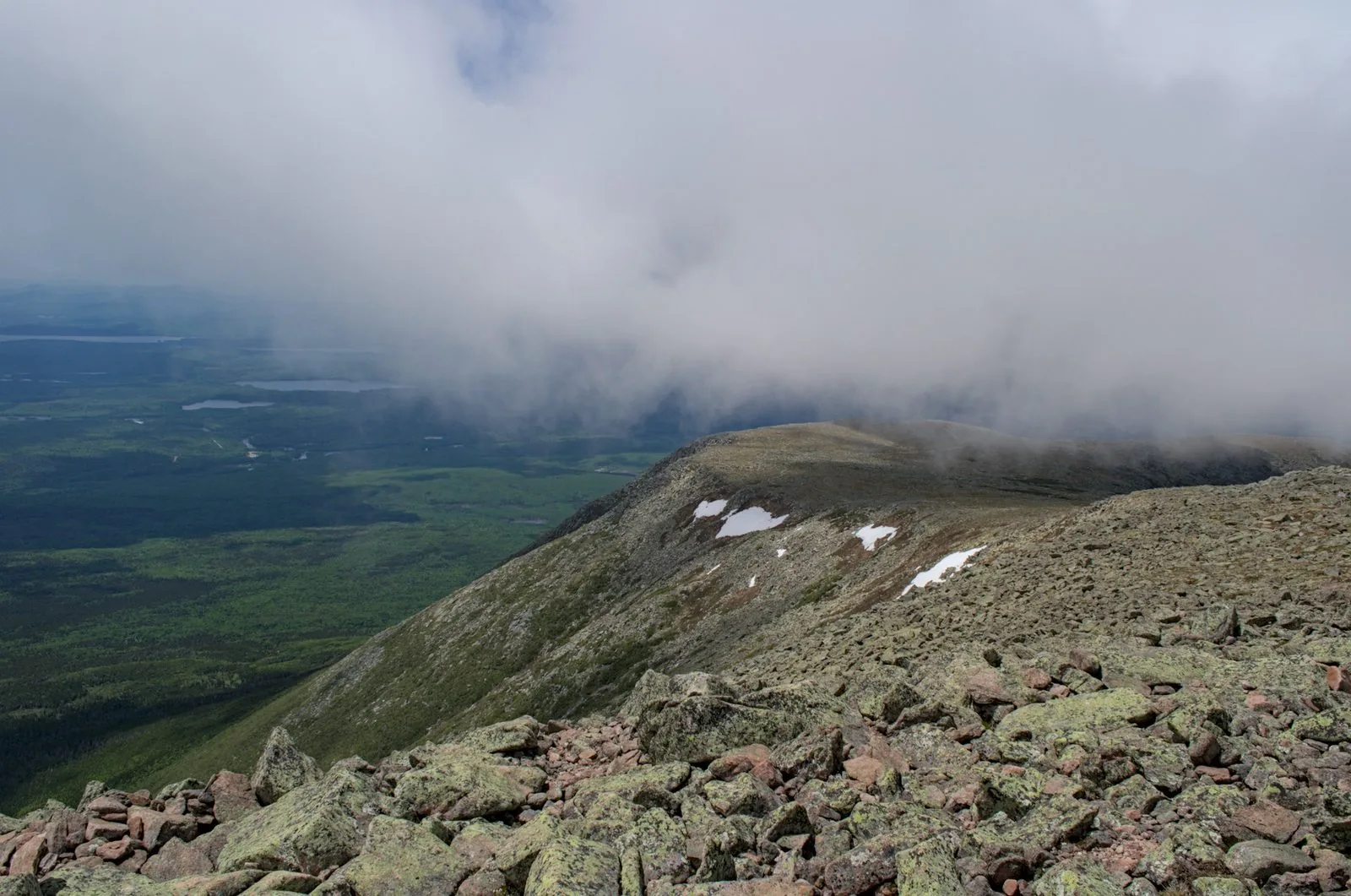 The Katahdin tableland with storm clouds rolling in from the west — patches of snow linger into summer, Baxter State Park wilderness stretching to the horizon below